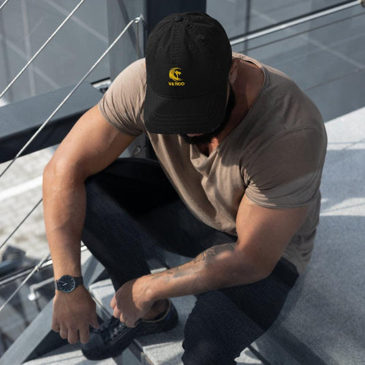 Man wearing a black U&TICO baseball cap and tan t-shirt sitting on outdoor stairs.